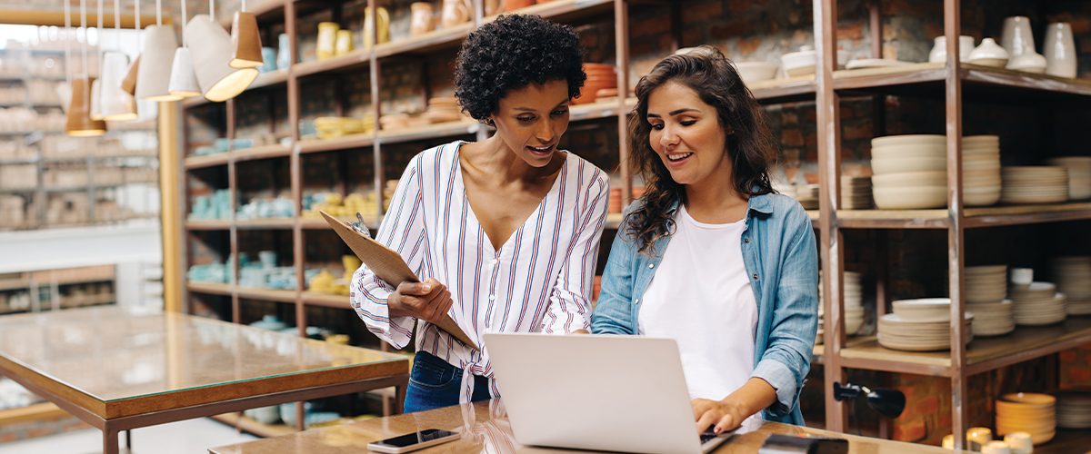 Two people reviewing information together on a laptop inside a retail shop with shelves of pottery and home goods, representing small‑business operations, inventory management, and collaborative workspace activity.